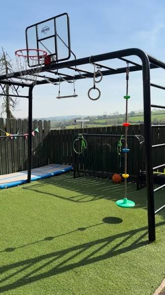 BERG PlayBase on artificial grass with a basketball hoop, trapeze bar and gym rings, with fields in the background