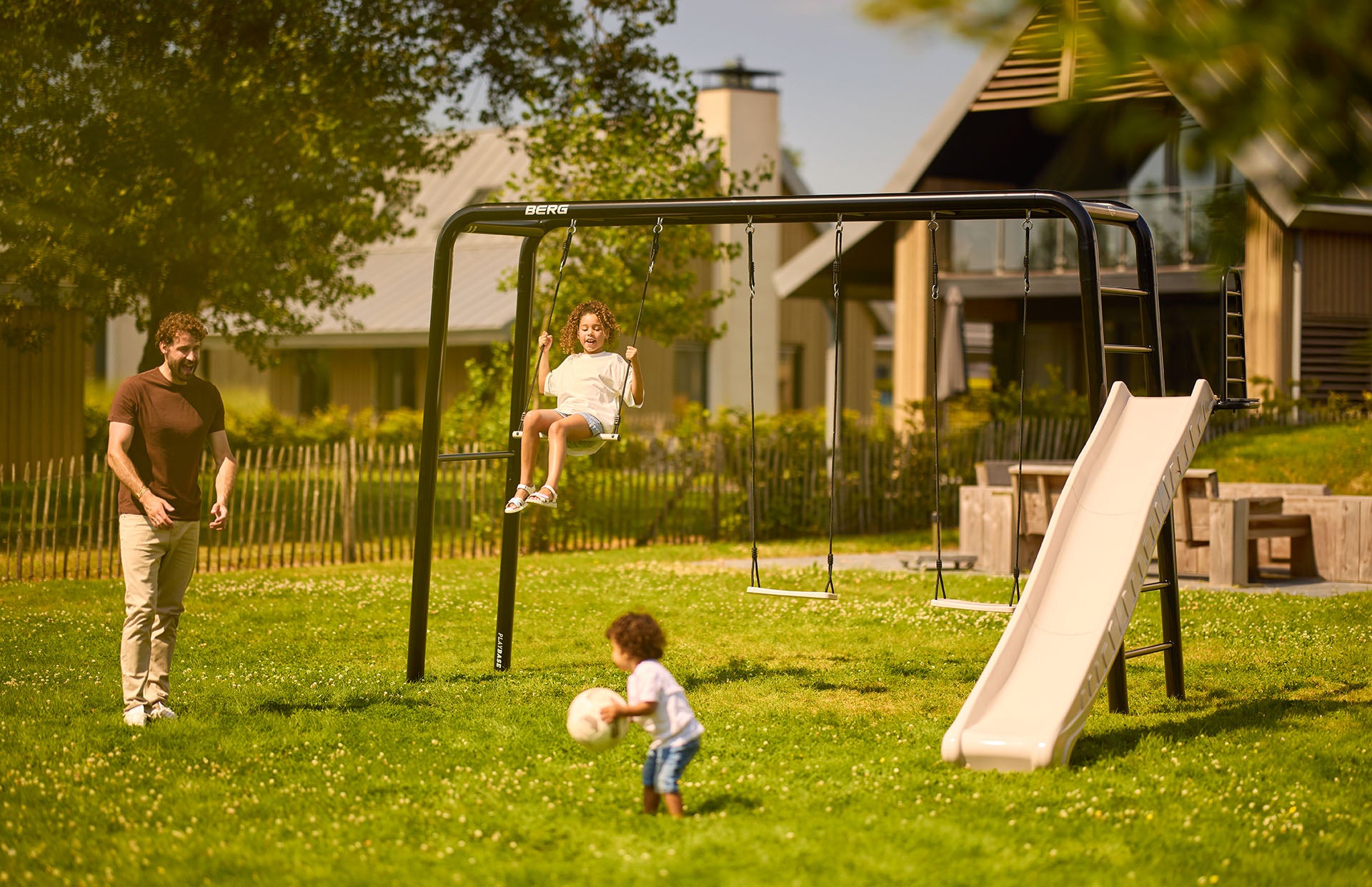 Dad and two children with a BERG PlayBase, swings and slide in their back garden